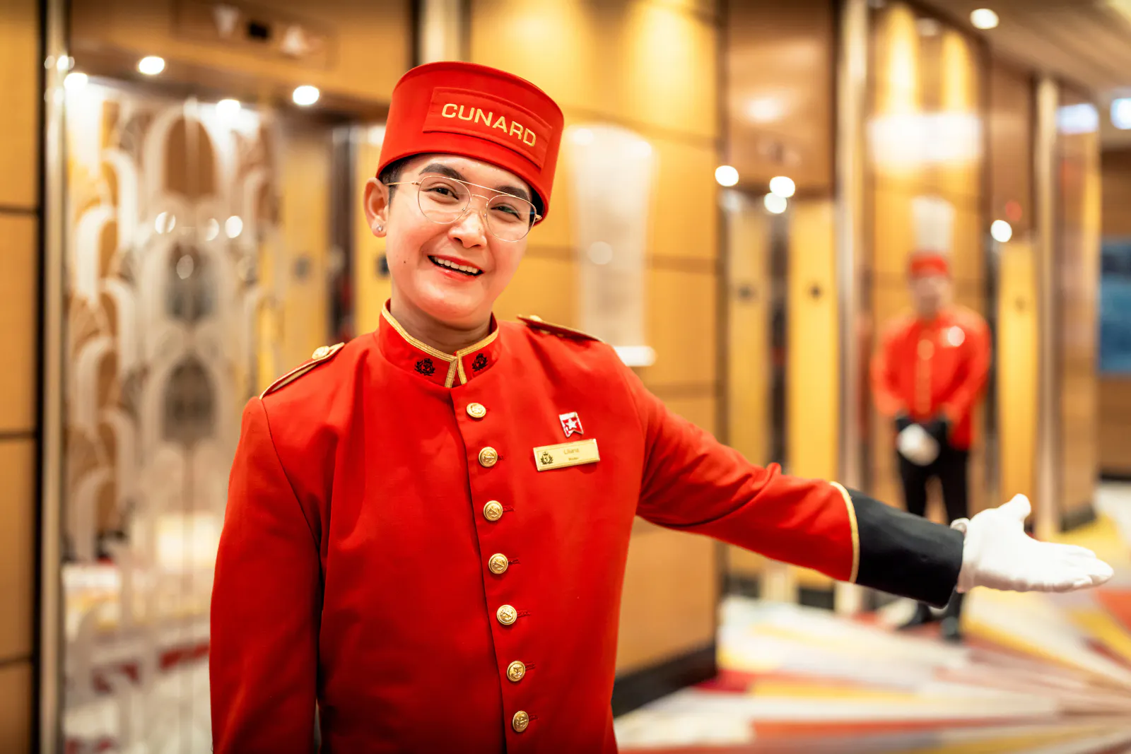 A smiling Cunard bellboy in a red uniform and cap welcomes guests on board Queen Victoria.