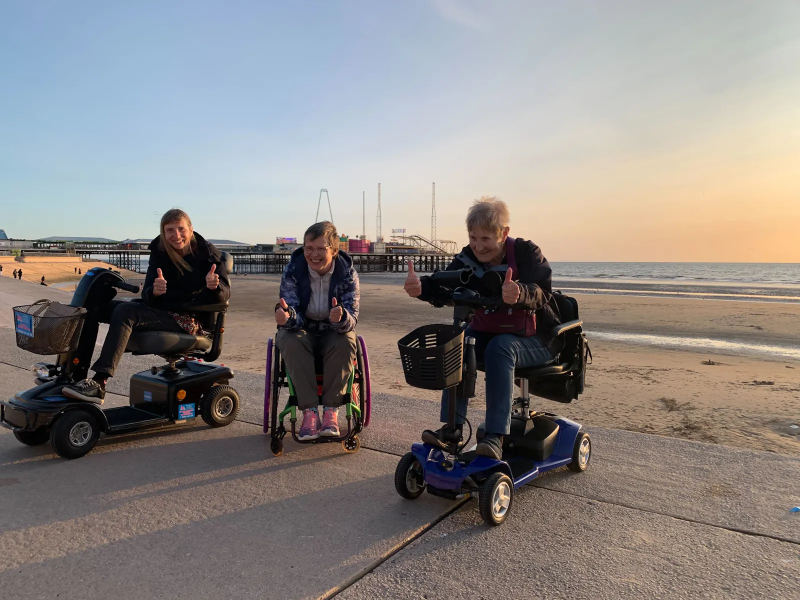 Guests using wheelchairs smiling in front of Blackpool pier as the sun sets.