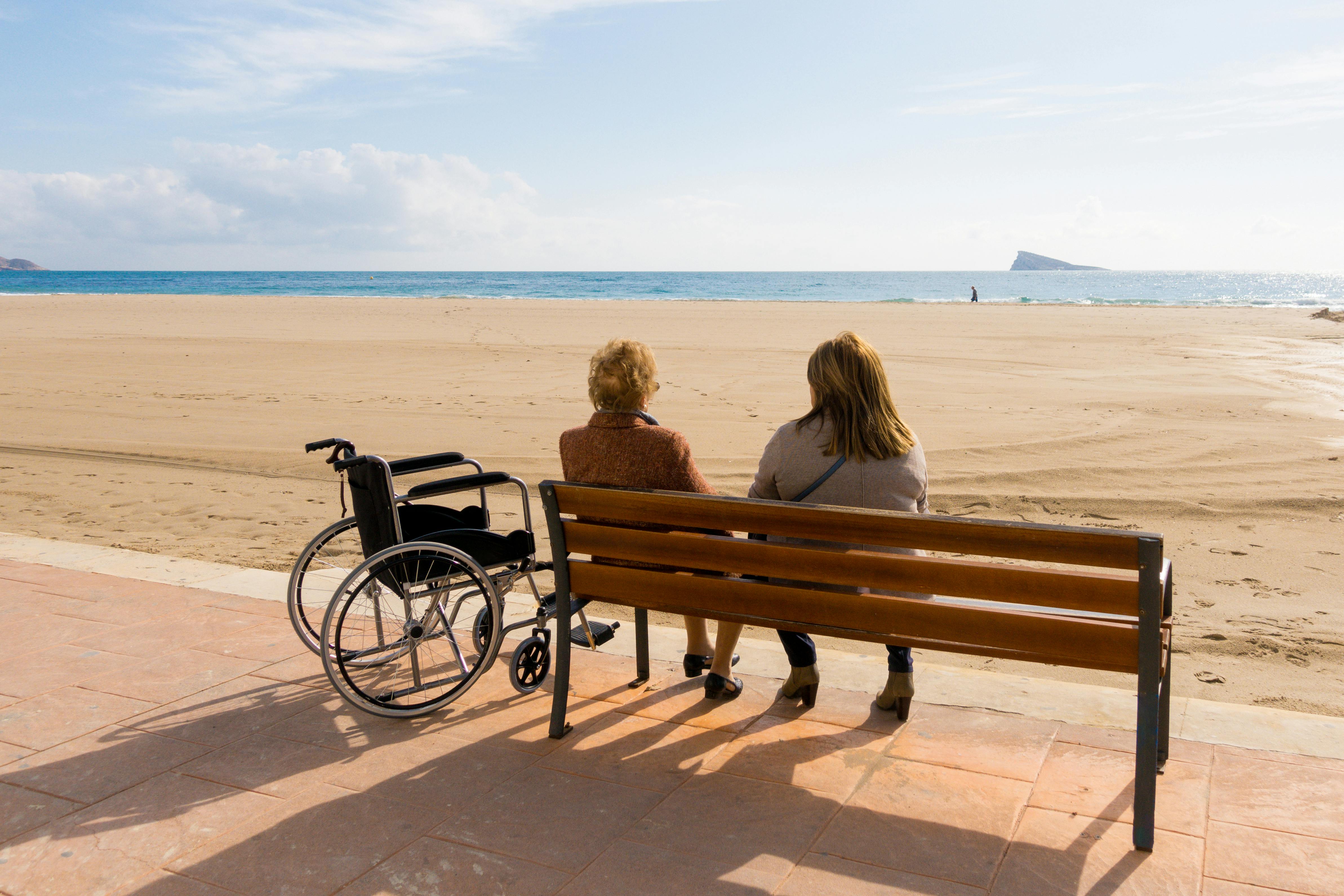 Two people sitting on a beach with a wheelchair nearby.
