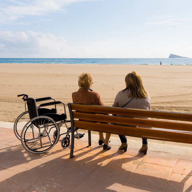 Two people sitting on a beach with a wheelchair nearby.