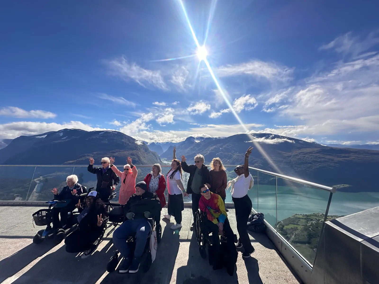 A group of Limitless guests, several using wheelchairs, raise their arms in celebration on a sunny viewing platform high above a fjord.