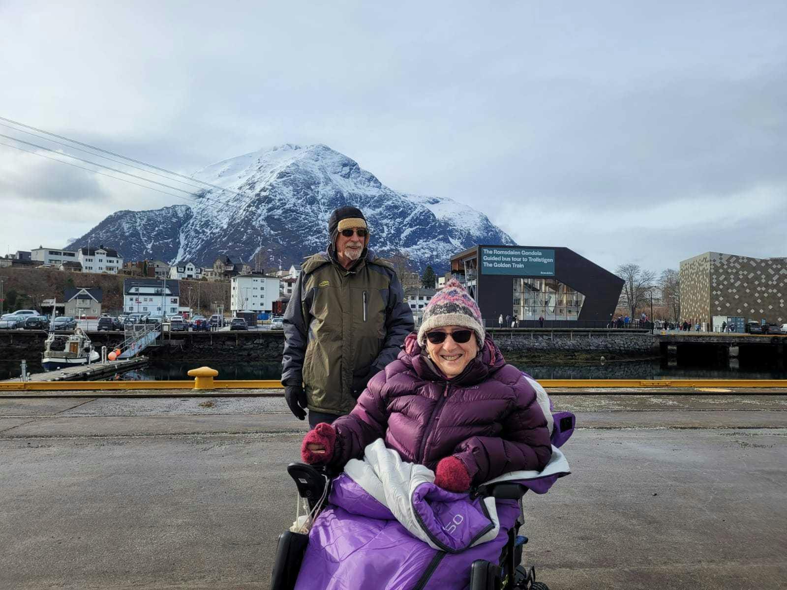 A smiling Limitless guest on a mobility scooter, wrapped up warm with a companion, on a Norwegian quayside beneath a snow-capped mountain.